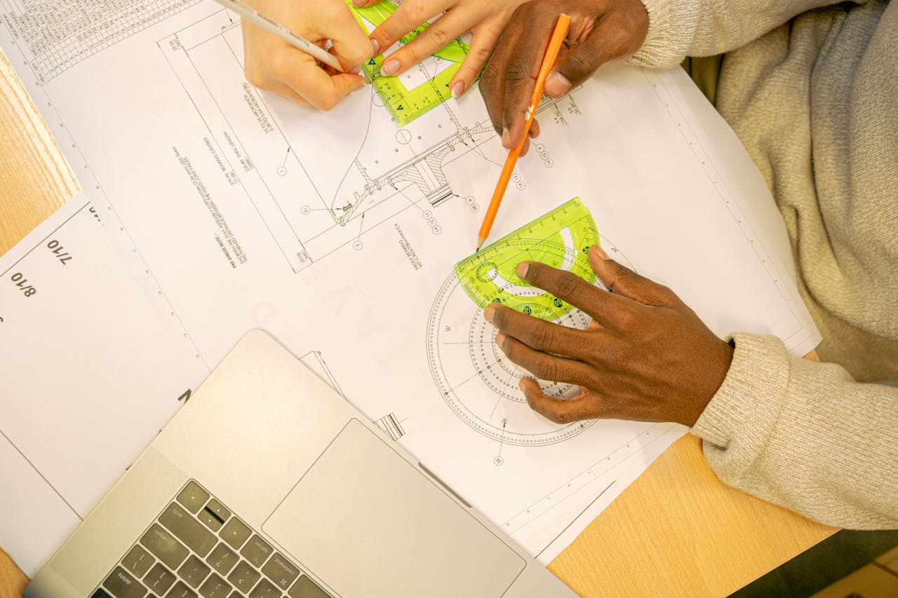 Hands collaborating on a blueprint drawing with a protractor and laptop, showcasing teamwork and design precision.